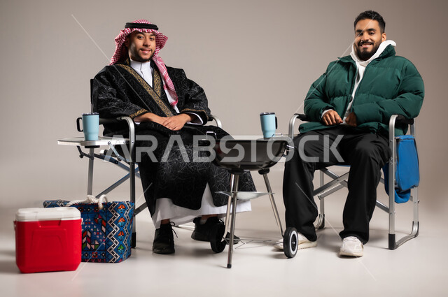 الاستمتاع بقضاء الوقت مع الأصدقاء , القيام بحفلات الشواء الممتعة , بورتريه لرجل عربي خليجي سعودي يرتدي الثوب التقليدي و الشماغ مع ملابس الشتاء الدافئة يجلس مع شاب بالزي الكاجوال , رحلات برية شبابية , خلفية رمادية