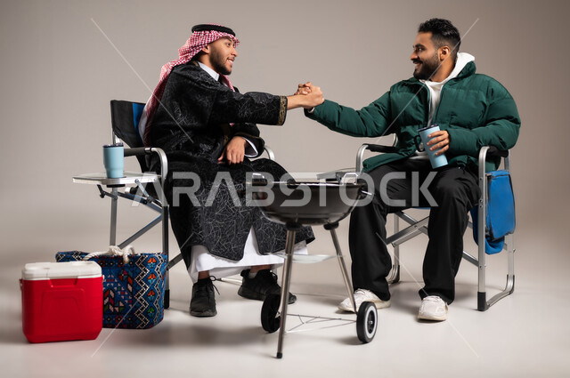 مفهوم الالفة و المودة بين الأصحاب , شوي اللحم على موقد الشواء , بورتريه لرجل عربي خليجي سعودي يرتدي الثوب التقليدي و الشماغ مع ملابس الشتاء الدافئة يجلس مع شاب بالزي الكاجوال , قضاء الأوقات الممتعة مع الأصدقاء , خلفية رمادية