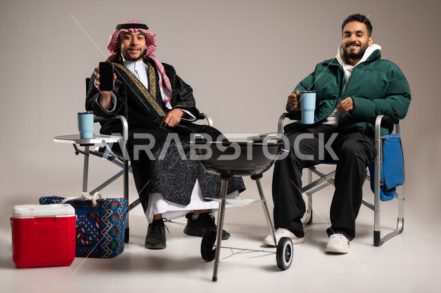 Looking at the camera with gestures of joy and pleasure, using technical devices to have fun with friends, portrait of a Saudi Gulf Arab man wearing a traditional thobe and shemagh with warm winter clothes sitting with a young man in casual clothes, an outdoor barbecue party, showing a blank black screen on a mobile phone, gray background