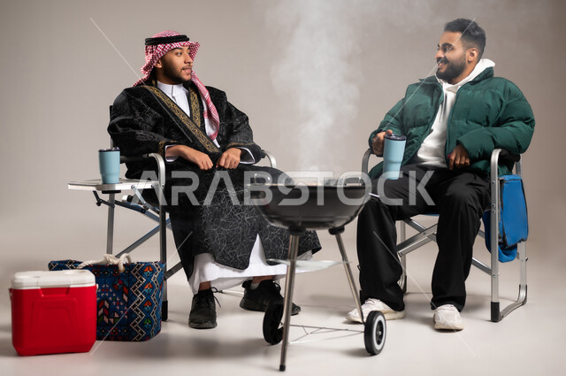 قضاء الأوقات الممتعة مع الأصدقاء , شوي اللحم على موقد الشواء , بورتريه لرجل عربي خليجي سعودي يرتدي الثوب التقليدي و الشماغ مع ملابس الشتاء الدافئة يجلس مع شاب بالزي الكاجوال , مفهوم الالفة و المودة بين الأصحاب , خلفية رمادية