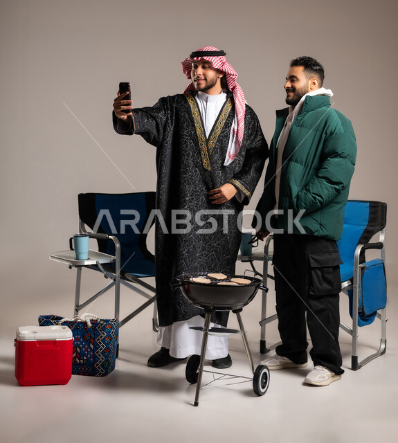 التقاط سيلفي تذكاري مع الأصدقاء , تحضير أطباق اللحوم المشوية على الفحم , بورتريه لرجل عربي خليجي سعودي يرتدي الثوب التقليدي و الشماغ مع ملابس الشتاء الدافئة يقف مع شاب بالزي الكاجوال , حفلة شواء في الهواء الطلق , صورة للجم بالطول الكامل , خلفية رمادية