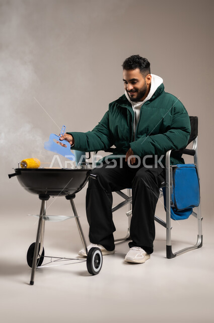 فن الطهي و الشواء , شوي الذرة الصفراء على الفحم , بورتريه لشاب عربي خليجي سعودي يرتدي الزي الكاجوال مع ملابس الشتاء الدافئة يجلس على كرسي الشواء يقوم بالتهوية لإحماء الفحم , صورة للجسم بالطول الكامل , خلفية رمادية