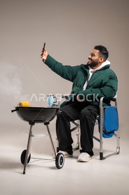 إجراء مكالمة الفيديو الهاتفية عبر الجوال , شوي الذرة الصفراء على الفحم , بورتريه لشاب عربي خليجي سعودي يرتدي الزي الكاجوال مع ملابس الشتاء الدافئة يمسك الهاتف المحمول بيده  , وجبات صحية مفيدة ذات قيمة غذائية عالية , صورة للجسم بالطول الكامل , خلفية رمادية