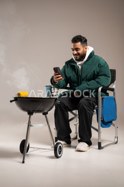 تصفح وسائل التواصل الاجتماعي عبر الجوال , شوي الذرة الصفراء على موقد الشواء , بورتريه لشاب عربي خليجي سعودي يرتدي الزي الكاجوال مع ملابس الشتاء الدافئة يمسك الهاتف المحمول بيده , رحلة برية ممتعة , صورة للجسم بالطول الكامل , خلفية رمادية
