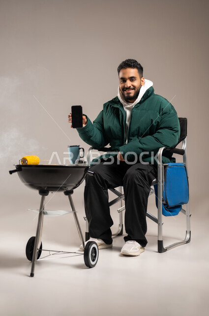 استخدام الهاتف المحمول للتسلية و الترفيه , شوي اللحم و الذرة الصفراء على موقد الشواء , بورتريه لشاب عربي خليجي سعودي يرتدي الزي الكاجوال مع ملابس الشتاء الدافئة , عرض شاشة سوداء فارغة عبر الجوال , صورة للجسم بالطول الكامل , خلفية رمادية
