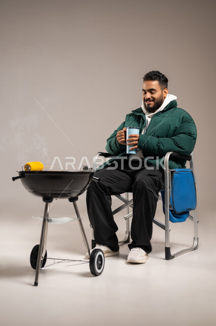 Enjoying hot drinks, a fun road trip, grilling meat and yellow corn on a barbecue stove, a portrait of a young Saudi Arabian Gulf Arab man wearing a casual outfit with warm winter clothes, a full-length body portrait, gray background
