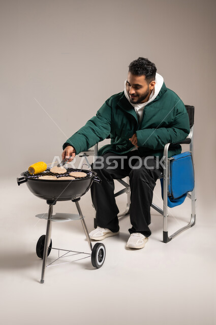 إعداد وجبات الطعام اللذيذة و الصحية , شوي اللحم و الذرة الصفراء على الفحم , بورتريه لشاب عربي خليجي سعودي يرتدي الزي الكاجوال مع ملابس الشتاء الدافئة يمسك الملقط بيديه , حفلة شواء ممتعة , صورة للجسم بالطول الكامل , خلفية رمادية