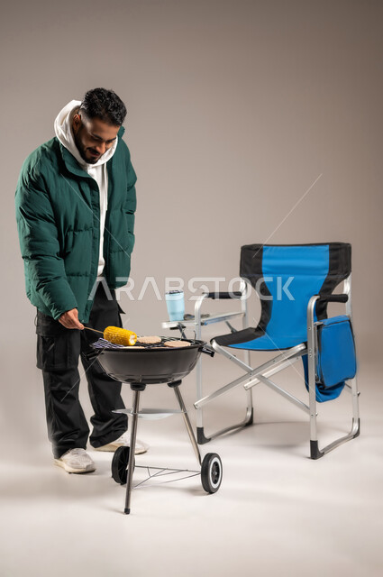 إعداد للحم و الذرة المشوية , رحلة برية ممتعة , بورتريه لشاب عربي خليجي سعودي يرتدي الزي الكاجوال مع ملابس الشتاء الدافئة يقوم بالشواء على الفحم , أطعمة و مأكولات شرقية , صورة للجسم بالطول الكامل , خلفية رمادية