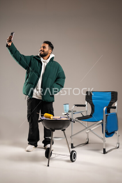 أحذ سيلفي تذكاري في الرحلة البرية , إعداد اللحم و الذرة المشوية على الطريقة الشرقية , بورتريه لشاب عربي خليجي سعودي يرتدي الزي الكاجوال مع ملابس الشتاء الدافئة يمسك الهاتف المحمول بيده , حفلة شواء في الهواء الطلق  , صورة للجسم بالطول الكامل , خلفية رمادية