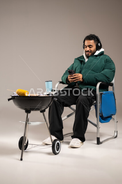 استخدام التقنيات الحديثة و المتطورة للتسلية , شوي اللحم و الذرة الصفراء على موقد الشواء , بورتريه لشاب عربي خليجي سعودي يرتدي الزي الكاجوال مع ملابس الشتاء الدافئة يضع سماعات الأذن الصوتية و يمسك الجوال بيده , صورة للجسم بالطول الكامل , خلفية رمادية