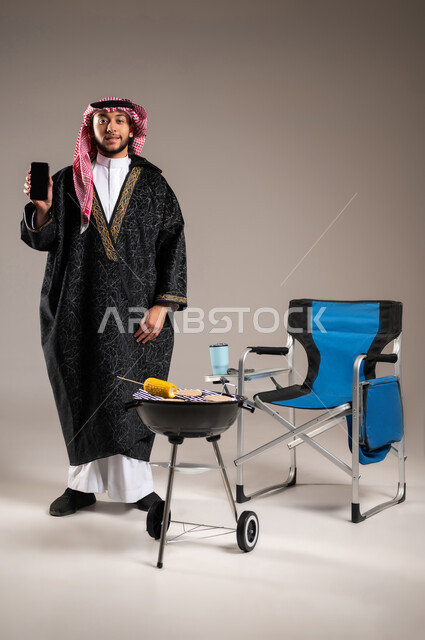 استخدام التكنولوجيا الحديثة و المتطورة , شوي اللحم و الذرة على الفحم , بورتريه لشاب عربي خليجي سعودي يرتدي الثوب التقليدي مع ملابس الشتاء الدافئة يقف أمام المنقل يمسك الهاتف المحمول بيده , وجبات صحية مفيدة ذات قيمة غذائية عالية ,صورة للجسم بالطول الكامل , خلفية رمادية