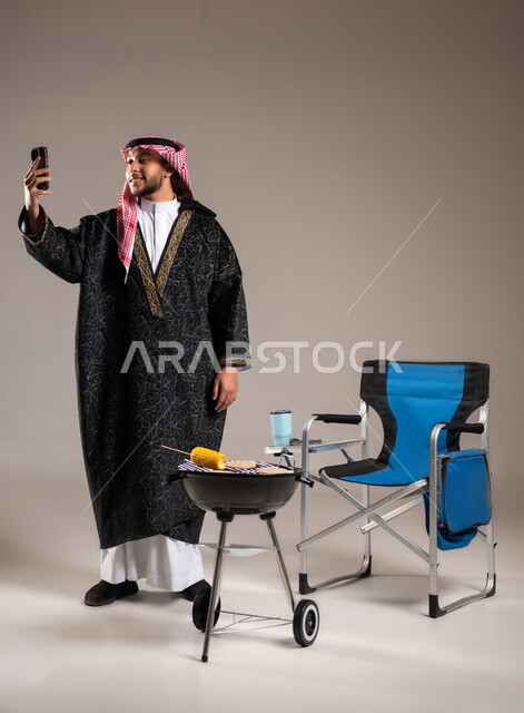 إجراء مكالمة فيديو هاتفية مع الأهل , شوي اللحم و الذرة على موقد الشواء , بورتريه لشاب عربي خليجي سعودي يرتدي الثوب التقليدي مع ملابس الشتاء الدافئة يقف أمام المنقل يمسك الهاتف المحمول بيده , رحلة برية ممتعة , صورة للجسم بالطول الكامل , خلفية رمادية
