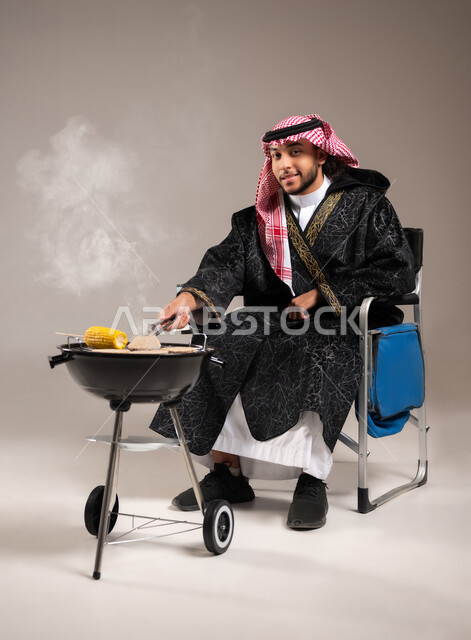 أصناف مشاوي شرقية , شوي الذرة الصفراء على موقد الشواء , بورتريه لشاب عربي خليجي سعودي يرتدي الثوب التقليدي و الشماغ يجلس على كرسي الشوي أمام المنقل يمسك الملقط بيده , إيماءات الفرح و السرور , صورة للجسم بالطول الكامل , خلفية رمادية