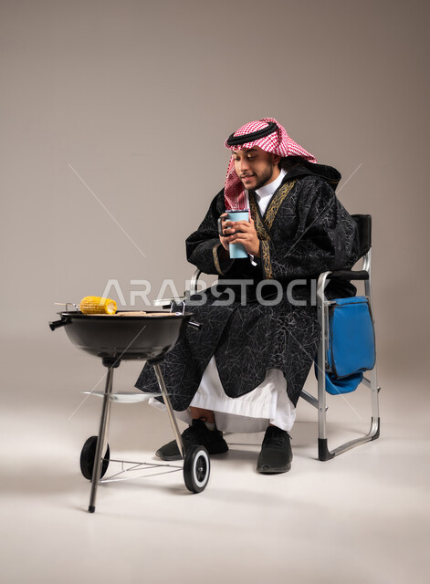 شرب المشروبات الشتوية الساخنة , رحلة برية ممتعة , بورتريه لشاب عربي خليجي سعودي يرتدي الثوب التقليدي و الشماغ يجلس على كرسي الشوي أمام المنقل , شوي الذرة الصفراء على موقد الشواء , صورة للجسم بالطول الكامل , خلفية رمادية