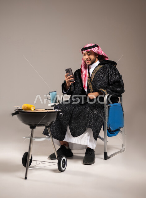 استخدام التكنولوجيا في التسلية و الترفيه , رحلة برية ممتعة , بورتريه لشاب عربي خليجي سعودي يرتدي الثوب التقليدي و الشماغ يجلس على كرسي الشوي أمام المنقل يمسك الهاتف المحمول بيده , حفلة شواء في الهواء الطلق , صورة للجسم بالكامل , خلفية رمادية