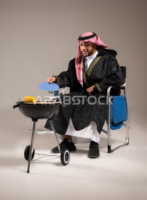 أصناف مشاوي شهية , شواء اللحم و الذرة على الفحم , بورتريه لشاب عربي خليجي سعودي يرتدي الثوب التقليدي و الشماغ مع ملابس الشتاء الدافئة يجلس على كرسي الشوي أمام المنقل يقوم بالتهوية لإحماء الفحم , صورة للجسم بالطول الكامل , خلفية رمادية