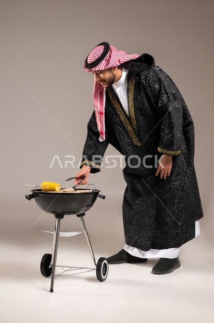 شواء اللحم و الذرة على الفحم , رحلة برية ممتعة , بورتريه لشاب عربي خليجي سعودي يرتدي الثوب التقليدي و الشماغ مع ملابس الشتاء الدافئة يقف أمام المنقل يمسك الملقط بيده , أصناف مشاوي شهية , صورة للجسم بالطول الكامل , خلفية رمادية