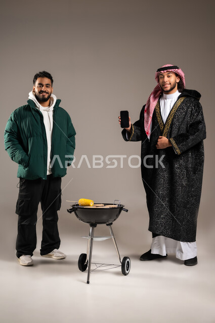 استخدام التقنيات الحديثة للترفيه , رحلة برية شبابية ممتعة , بورتريه لشاب عربي خليجي سعودي يرتدي الثوب التقليدي و الشماغ مع ملابس الشتاء الدافئة يقف مع شاب بالزي الكاجوال , حفلة شواء مع الأصدقاء , صورة للجسم بالطول الكامل , خلفية رمادية