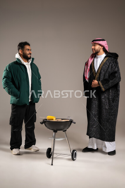 مفهوم الالفة و المحبة بين الأصدقاء , رحلة برية مع الأصحاب , بورتريه لشاب عربي خليجي سعودي يرتدي الثوب التقليدي و الشماغ مع ملابس الشتاء الدافئة يقف مع شاب بالزي الكاجوال , حفلة شواء في الهواء الطلق , صورة للجسم بالطول الكامل , خلفية رمادية