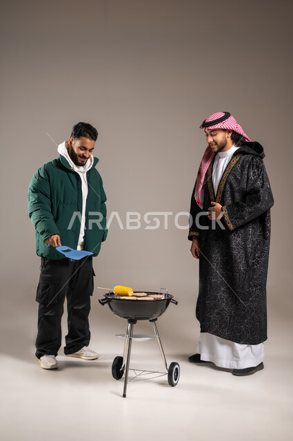حفلة شواء مع الأصدقاء , إعداد اللحوم و الذرة المشوية , بورتريه لشاب عربي خليجي سعودي يرتدي الثوب التقليدي و الشماغ مع ملابس الشتاء الدافئة يقف مع شاب بالزي الكاجوال يقوم بالتهوية لإحماء الفحم  , صورة للجسم بالطول الكامل , خلفية رمادية