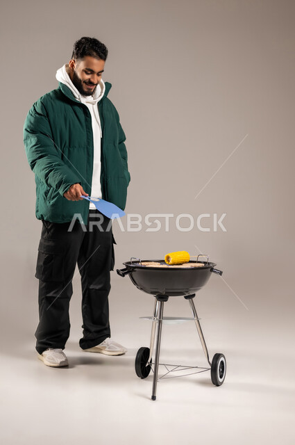 رحلة برية ممتعة , وجبات صحية مفيدة ذات قيمة غذائية عالية  , شوي الذرة الصفراء على موقد الشواء , بورتريه لشاب عربي خليجي سعودي يرتدي الزي الكاجوال مع ملابس الشتاء الدافئة يقف أمام المنقل يقوم بالتهوية لإحماء الفحم , صورة للجسم بالطول الكامل , خلفية رمادية