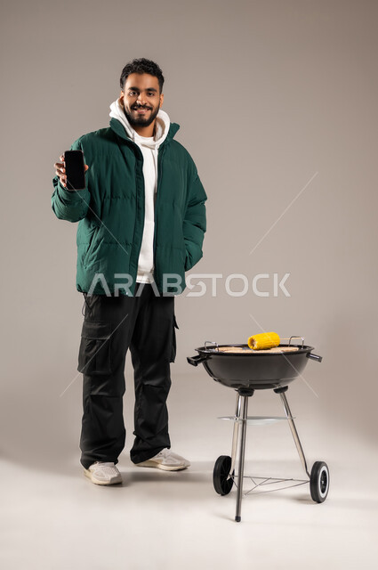 استخدام لأجهزة التقنية للتسلية و الترفيه , شوي الذرة الصفراء على موقد الشواء , بورتريه لشاب عربي خليجي سعودي يرتدي الزي الكاجوال مع ملابس الشتاء الدافئة يقف أمام المنقل يمسك الجوال بيده , عرض شاشة سوداء فارغة عبر الهاتف المحمول , صورة للجسم بالطول الكامل , خلفية رمادية