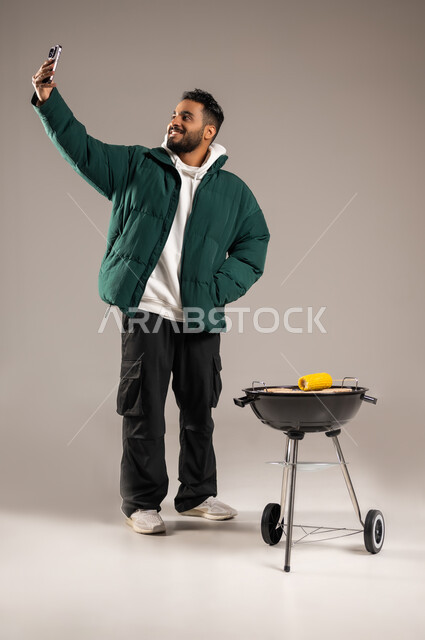 استخدام الهاتف المحمول في التقاط الصور , رحلة برية ممتعة , بورتريه لشاب عربي خليجي سعودي يرتدي الزي الكاجوال مع ملابس الشتاء الدافئة يقف أمام المنقل يمسك الجوال بيده , شوي الذرة على الفحم , صورة للجسم بالطول الكامل , خلفية رمادية
