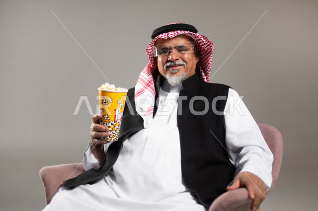 إحضار السناكات اللذيذة والاستعداد لمشاهدة الافلام، مشاهدة فلم سينمائي، بورتريه مقرب لرجل عربي خليجي سعودي مبتسم كبير بالسن يرتدي الزي التقليدي و الشماغ يحمل بيده علبة الفوشار، إيماءات السعادة والمتعة، خلفية رمادية