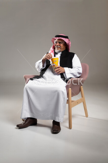 الاستعداد لمشاهدة الافلام وإحضار السناكات اللذيذة، مشاهدة فلم سينمائي، بورتريه لرجل عربي خليجي سعودي مبتسم كبير بالسن يرتدي الزي التقليدي و الشماغ يحمل بيده علبة الفوشار، إيماءات السعادة والمتعة، خلفية رمادية
