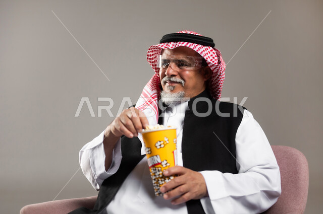إحضار السناكات اللذيذة والاستعداد لمشاهدة الافلام، مشاهدة فلم سينمائي، بورتريه مقرب لرجل عربي خليجي سعودي مبتسم كبير بالسن يرتدي الزي التقليدي و الشماغ يحمل بيده علبة الفوشار، إيماءات السعادة والمتعة، خلفية رمادية
