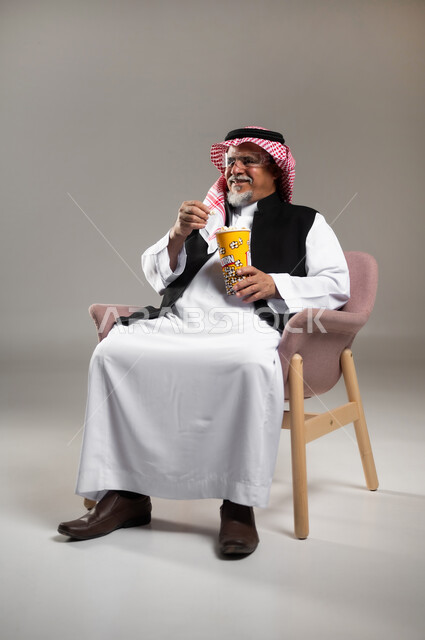 الاستمتاع بمشاهدة فلم سينمائي، قضاء أوقات ممتعة ومسلية، بورتريه لرجل عربي خليجي سعودي مبتسم كبير بالسن يرتدي الزي التقليدي و الشماغ يحمل بيده علبة الفوشار يستمتع بمشاهدة الأفلام ، إيماءات السعادة والمتعة، خلفية رمادية