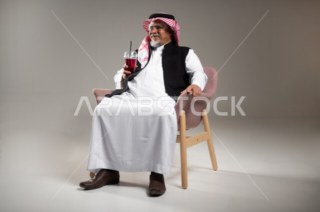 الاستمتاع بتناول المشروبات المفيدة اللذيذة، بورتريه مقرب لرجل كبير بالسن عربي خليجي سعودي مبتسم يرتدي الثوب التقليدي والشماغ يستمتع بشرب العصير الطبيعي البارد، محاولة التخفيف من حرارة الصيف بالمشروب المنعش، خلفية رمادية