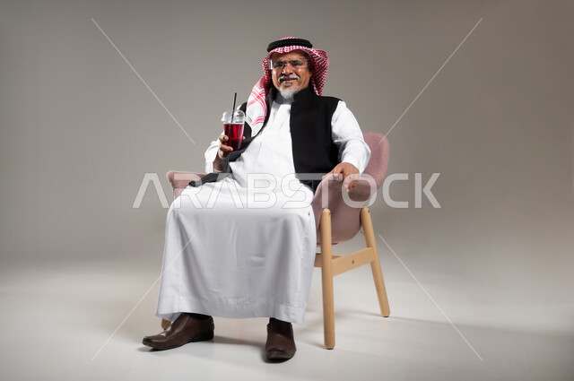 Enjoying a delicious healthy drink, close-up portrait of a smiling elderly Saudi Arabian Gulf man wearing traditional thobe and shemagh enjoying drinking cold natural juice, trying to cool down with the summer heat with a refreshing drink, gray background