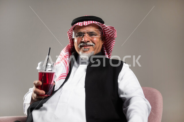 محاولة التخفيف من حرارة الصيف بالمشروب المنعش، الاستمتاع بتناول المشروبات المفيدة اللذيذة، بورتريه مقرب لرجل كبير بالسن عربي خليجي سعودي مبتسم يرتدي الثوب التقليدي والشماغ يستمتع بشرب العصير الطبيعي البارد، خلفية رمادية
