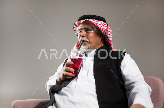 محاولة التخفيف من حرارة الصيف بالمشروب المنعش، الاستمتاع بتناول المشروبات المفيدة اللذيذة، بورتريه مقرب لرجل كبير بالسن عربي خليجي سعودي مبتسم يرتدي الثوب التقليدي والشماغ يستمتع بشرب العصير الطبيعي البارد، خلفية رمادية