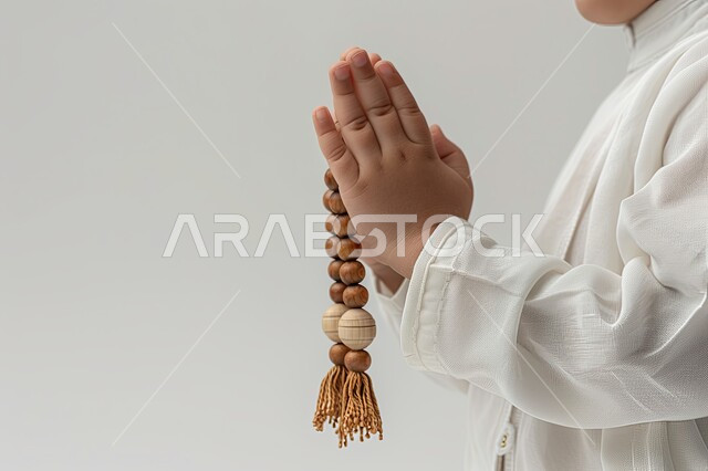 الإكثار من ذكر الله تعالى والإستغفار والدعاء, مفهوم الإجتهاد في العباده و التقرب الى الله في شهر رمضان المبارك, صورة مقربة لأيدي ولد عربي سعودي خليجي يرتدي الثوب الأبيض يرفع يديه يمسك السبحه, يوم الجمعه المباركه، خلفيه بيضاء