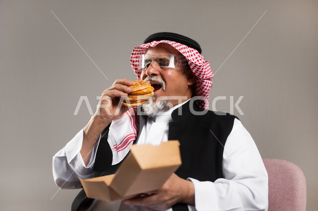 مساوئ الإكثار من أكل الوجبات السريعة للكبار بالعمر, الاستمتاع بتناول الطعام الجاهز, بورتريه مقرب لرجل عربي خليجي سعودي كبير في السن يرتدي الثوب التقليدي و الشماغ يجلس على الكرسي يقوم بأكل البرغر, خلفية رمادية