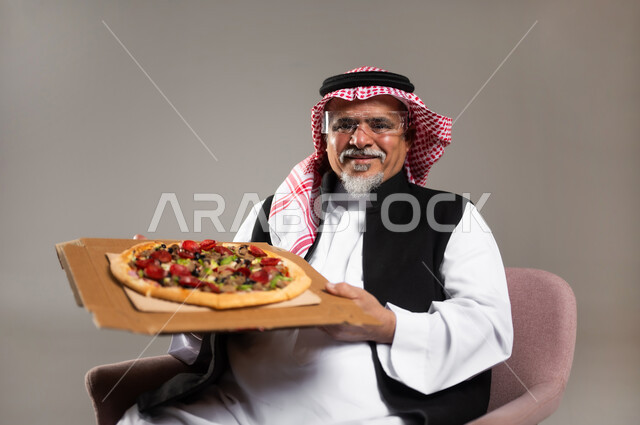 أضرار تناول المأكولات السريعة على الكبار بالعمر، تناول المخبوزات والمعجنات, مفهوم وجبات الطعام غير المفيدة, بورتريه مقرب لرجل كبير بالسن عربي خليجي سعودي مبتسم يرتدي الثوب التقليدي و الشماغ يجلس على الكرسي و يحمل بيده البيتزا, خلفية رمادية