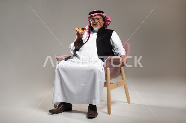 تناول المخبوزات والمعجنات الطازجة, مفهوم وجبات الطعام غير المفيدة, بورتريه لرجل كبير بالسن عربي خليجي سعودي مبتسم يرتدي الثوب التقليدي و الشماغ يجلس على الكرسي و يحمل بيده قطعة من البيتزا, أضرار تناول المأكولات السريعة على الكبار بالعمر، خلفية رمادية