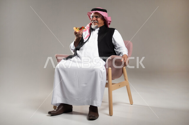 تناول المخبوزات والمعجنات الطازجة, مفهوم وجبات الطعام غير المفيدة, بورتريه لرجل كبير بالسن عربي خليجي سعودي مبتسم يرتدي الثوب التقليدي و الشماغ يجلس على الكرسي و يحمل بيده قطعة من البيتزا, أضرار تناول المأكولات السريعة على الكبار بالعمر، خلفية رمادية