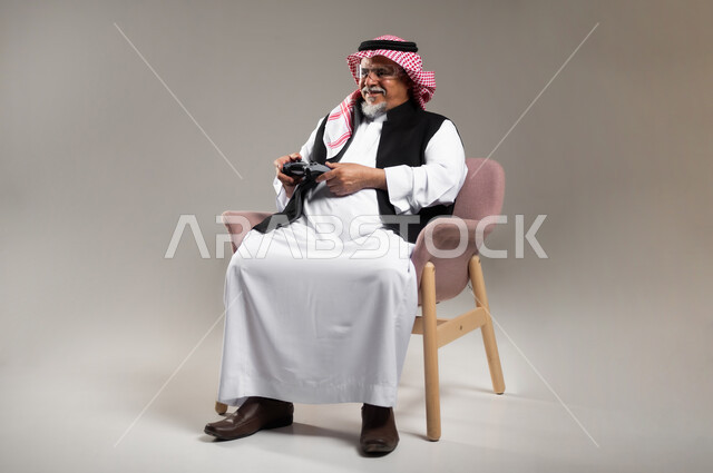 قضاء وقت ممتع بالألعاب الالكترونية، تعابير التحدي و المغامرة, بورتريه لرجل عربي خليجي سعودي مبتسم كبير بالسن يرتدي الزي التقليدي و الشماغ يمسك بيده عصا التحكم بألعاب الفيديو بإيماءات الحماس, خلفية رمادية