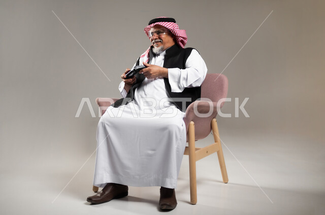 تعابير التحدي و المغامرة, قضاء وقت ممتع بالألعاب الالكترونية، بورتريه لرجل عربي خليجي سعودي مبتسم كبير بالسن يرتدي الزي التقليدي و الشماغ يمسك بيده عصا التحكم بألعاب الفيديو بإيماءات الحماس, خلفية رمادية