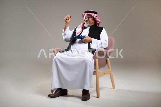 قضاء وقت ممتع بالألعاب الالكترونية، بورتريه لرجل عربي خليجي سعودي مبتسم كبير بالسن يرتدي الزي التقليدي و الشماغ يمسك بيده عصا التحكم بألعاب الفيديو يشير بيده للاعلى بتعبير الفوز, خلفية رمادية