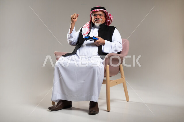 المتعة و التسلية بالألعاب الالكترونية، بورتريه لرجل عربي خليجي سعودي مبتسم كبير بالسن يرتدي الزي التقليدي و الشماغ يمسك بيده عصا التحكم بألعاب الفيديو, النظر الى الكاميرا بتعابير الفرح بالفوز, خلفية رمادية