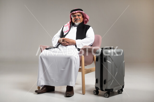 Waiting for the time of the tourist trip with gestures of joy and pleasure, searching and exploring around the world, portrait of an old Saudi Gulf Arab man wearing the traditional Saudi dress and shemagh sitting on the chair with clasped hands next to the luggage bag, gray background