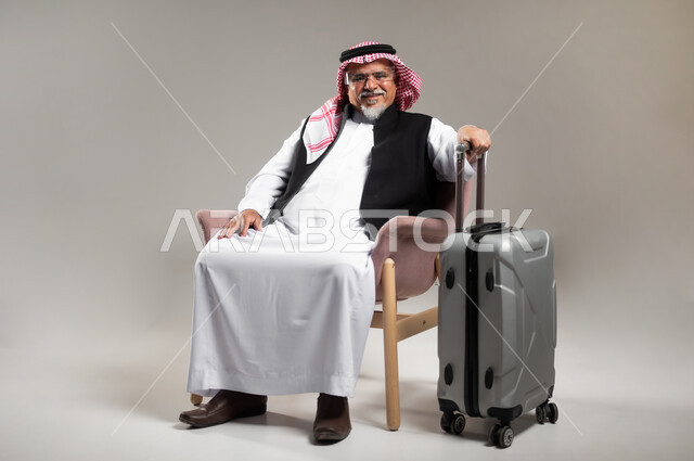 الرحلات السياحيه الممتعه، مفهوم البحث و الاستكشاف حول العالم, بورتريه لرجل عربي خليجي سعودي كبير بالسن يرتدي الزي السعودي التقليدي والشماغ يجلس على الكرسي و يمسك بيده حقيبة الامتعة, النظر الى الكاميرا بايماءات الفرح و السرور, خلفيه رماديه