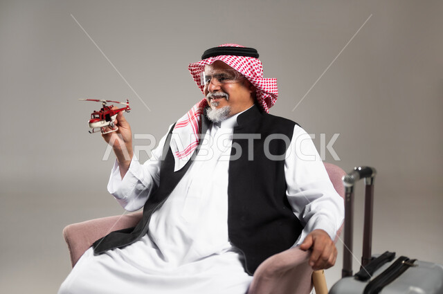 مفهوم الرحلات الجويه الترفيهيه، الاستعداد لقضاء وقت ممتع في السفر حول العالم, بورتريه مقرب لرجل عربي خليجي سعودي مبتسم كبير بالسن يرتدي الثوب التقليدي والشماغ يجلس بجانب حقيبة الامتعة ويحمل بيده مجسم طائرة حمراء, تعابير وايماءات الفرح و السرور, خلفية رمادية
