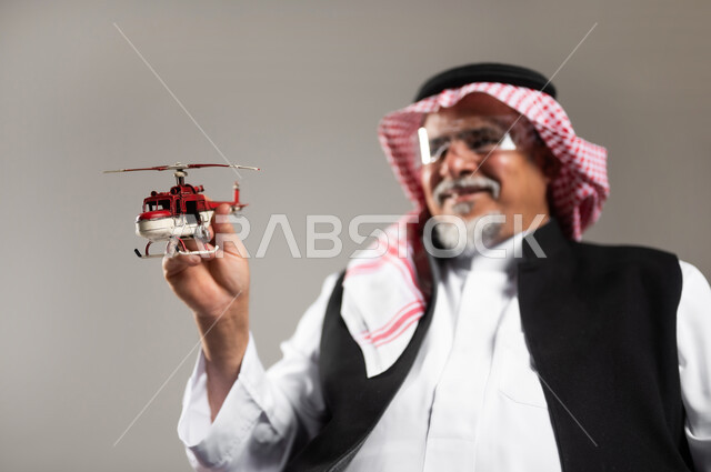 تعابير وايماءات الفرح و السرور, مفهوم الرحلات الجويه الترفيهيه، الاستعداد لقضاء وقت ممتع في السفر حول العالم, بورتريه مقرب لرجل عربي خليجي سعودي مبتسم كبير بالسن يرتدي الثوب التقليدي والشماغ يحمل بيده مجسم طائرة حمراء, خلفية رمادية