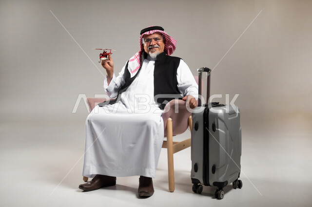 الاستعداد لقضاء وقت ممتع في السفر حول العالم, بورتريه لرجل عربي خليجي سعودي مبتسم كبير بالسن يرتدي الثوب التقليدي والشماغ يجلس بجانب حقيبة الامتعة ويحمل بيده مجسم طائرة حمراء, تعابير وايماءات الفرح و السرور, خلفية رمادية
