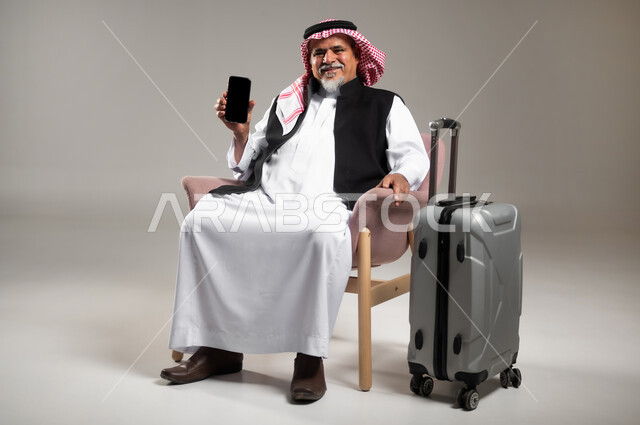 الحجوزات الالكترونية للرحلات السياحية, الاستعداد لقضاء وقت ممتع في العطلة، بورتريه لرجل عربي خليجي سعودي مبتسم كبير بالسن يرتدي الثوب التقليدي والشماغ يجلس على الكرسي بجانب حقيبة الامتعة يحمل بيده الهاتف الجوال بشاشة سوداء فارغة, خلفية رمادية